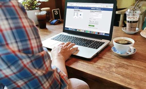 Man on the Facebook login page on his laptop, on a wooden table with a coffee next to the laptop