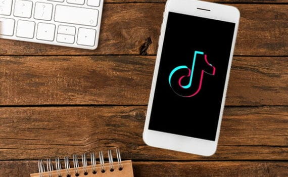 Mobile phone with the TikTok logo on the screen lying on a wooden table, with the corner of a white keyboard and top of a small spiral notepad visible