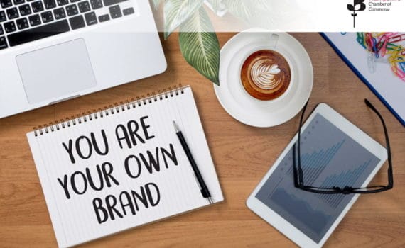 A spiral notebook with the words 'You are your own brand' written on it, lying next to a laptop kyeboard, tablet, and a cup of coffee, with some colourful paper clips in the top right corner