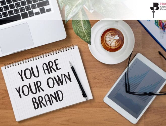 A spiral notebook with the words 'You are your own brand' written on it, lying next to a laptop kyeboard, tablet, and a cup of coffee, with some colourful paper clips in the top right corner