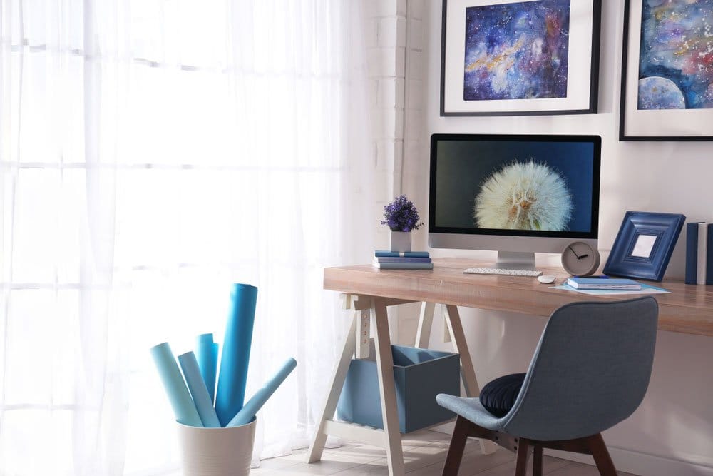 A desk by a bright window, with a plant on it, and framed pictures on the wall in front of it - creative space to work from home
