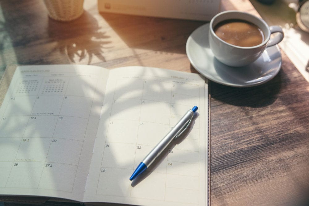 wooden desk with a day planner diary and a cup of tea next to it - plannign your day for working from home