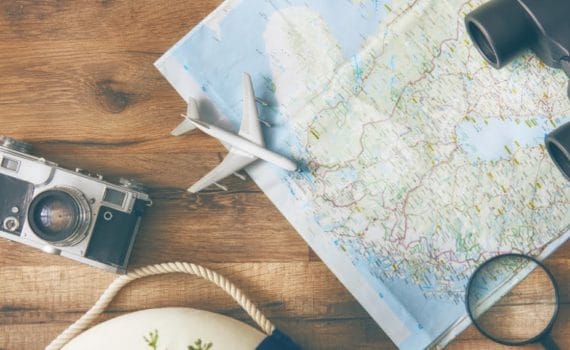 A map, camera, pair of binoculars, and a toy aeroplane sit on a table representing travel marketing.