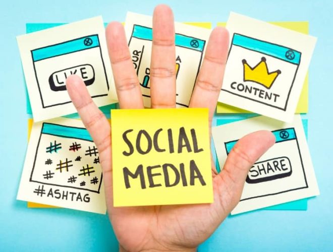 Hand holding sticky note with “Social Media” over background notes for likes, content, and hashtags, representing social media marketing planning.