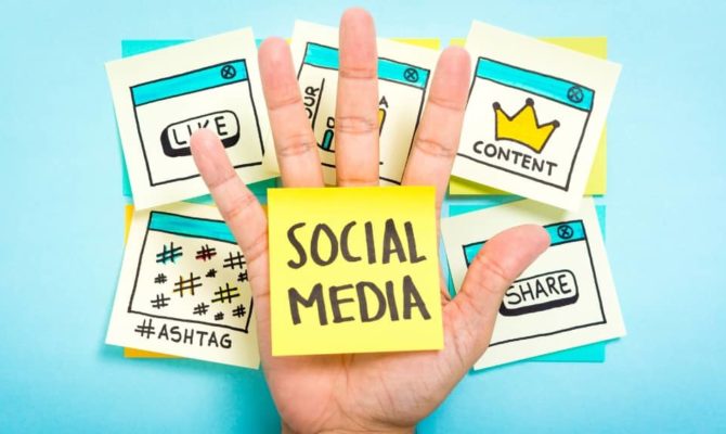 Hand holding sticky note with “Social Media” over background notes for likes, content, and hashtags, representing social media marketing planning.