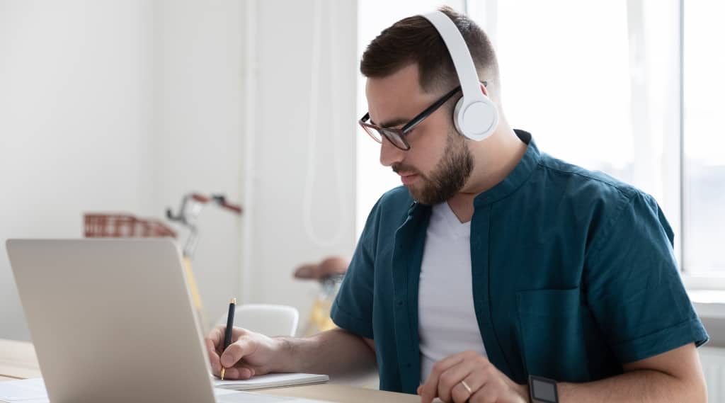 Man with headphones working on laptop and taking notes