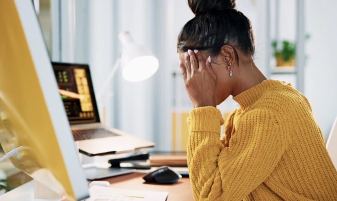 bad website design reaction, a frustrated woman in a yellow jumper holding her head in her hands at a desk with her computer