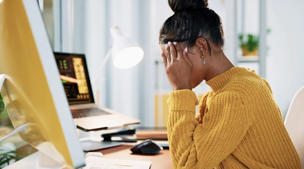 bad website design reaction, a frustrated woman in a yellow jumper holding her head in her hands at a desk with her computer