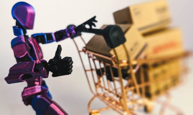 How AI Shopping Agents Choose Products for Black Friday: A purple and blue jointed robot figure gives a thumbs up while pushing a miniature copper shopping trolley loaded with cardboard parcels.