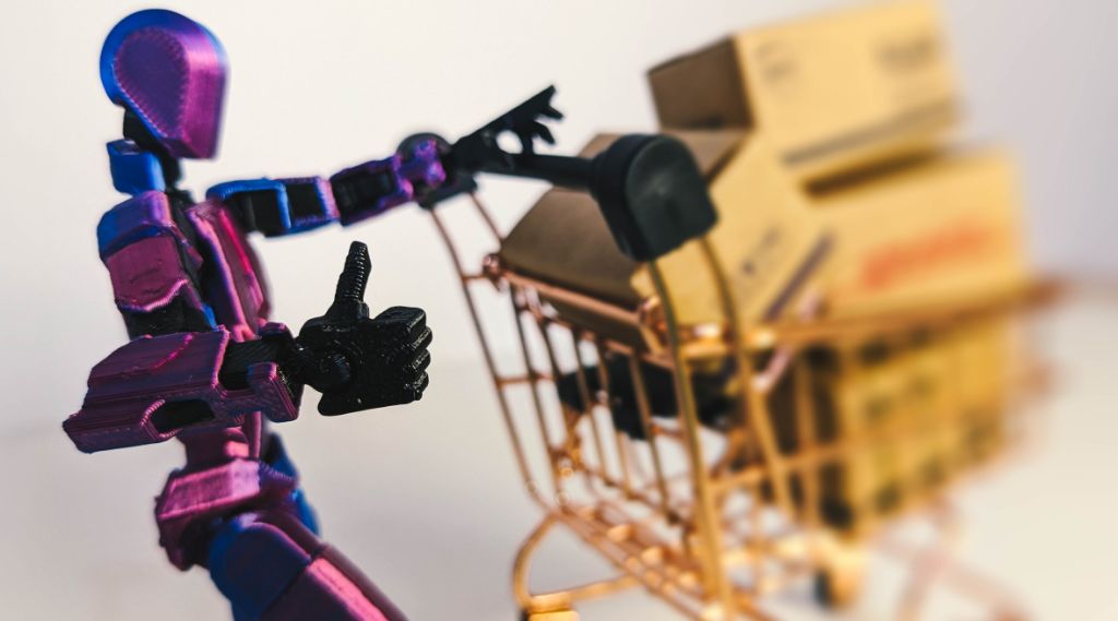 How AI Shopping Agents Choose Products for Black Friday: A purple and blue jointed robot figure gives a thumbs up while pushing a miniature copper shopping trolley loaded with cardboard parcels.