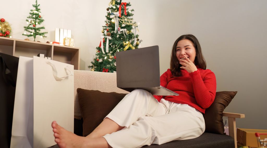 best time to publish Christmas gift guides: A smiling young woman in a red jumper and white trousers sits on a sofa with a laptop on her lap. Around her are a decorated Christmas tree, shopping bags and a small parcel.
