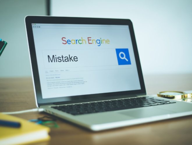 common SEO mistakes: A laptop on a wooden desk showing a search engine with the word "Mistake" typed into the search bar, beside a magnifying glass resting on printed SEO reports and a pot of coloured pens.