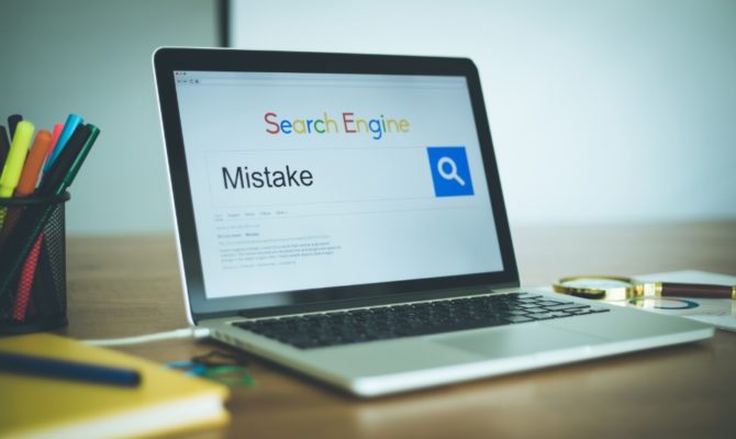 common SEO mistakes: A laptop on a wooden desk showing a search engine with the word "Mistake" typed into the search bar, beside a magnifying glass resting on printed SEO reports and a pot of coloured pens.