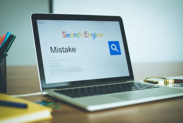 common SEO mistakes: A laptop on a wooden desk showing a search engine with the word "Mistake" typed into the search bar, beside a magnifying glass resting on printed SEO reports and a pot of coloured pens.