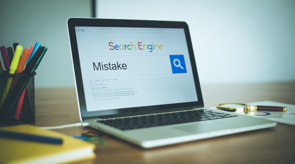 common SEO mistakes: A laptop on a wooden desk showing a search engine with the word "Mistake" typed into the search bar, beside a magnifying glass resting on printed SEO reports and a pot of coloured pens.