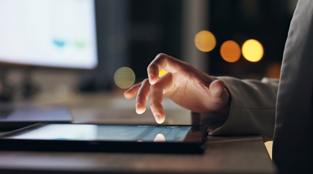 does my business need a website: A business owner's hand tapping a tablet screen at night, with a computer monitor and warm bokeh lights in the background, representing customers searching for businesses online at any hour.