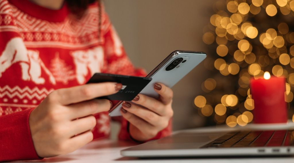 how far in advance to plan Christmas marketing: A person in a red and white festive jumper holds a bank card and a smartphone over a laptop. A lit red candle and the blurred golden lights of a Christmas tree glow in the background.