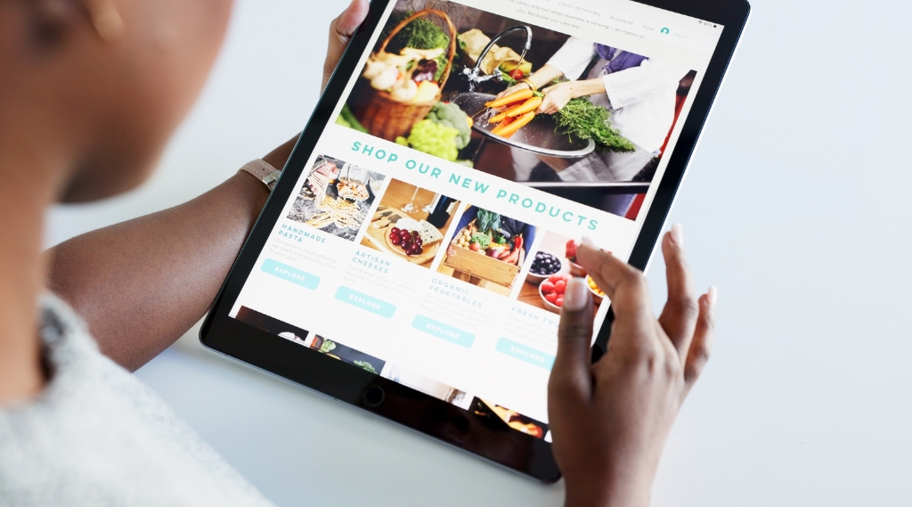what types of business need a website the mos: A woman holding a tablet showing a food retailer's website with the heading "Shop Our New Products" and categories for handmade pasta, artisan cheeses and organic vegetables, illustrating the type of business that benefits most from a website.